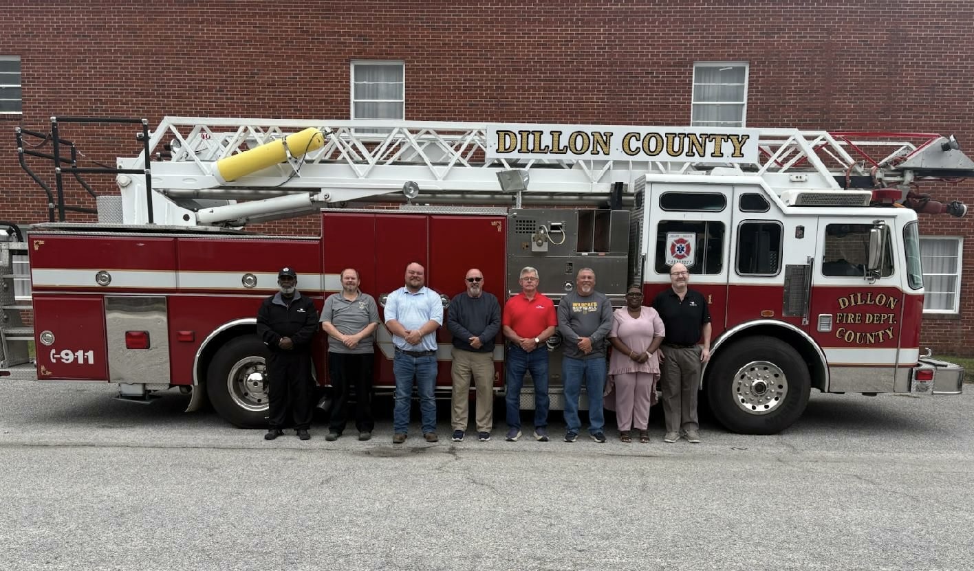A group of people stands in front of a red and white Dillon County fire truck parked beside a brick building.