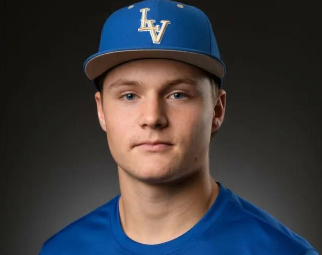 Young man wearing a blue baseball cap with "LV" letters and a matching blue shirt, looking directly at the camera with a neutral expression against a dark background.