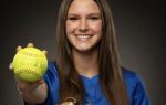 Smiling girl in blue jersey holding a softball with "2026" written on it, wearing a glove, against a dark background.