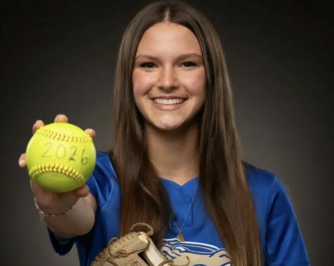 Smiling girl in blue jersey holding a softball with "2026" written on it, wearing a glove, against a dark background.