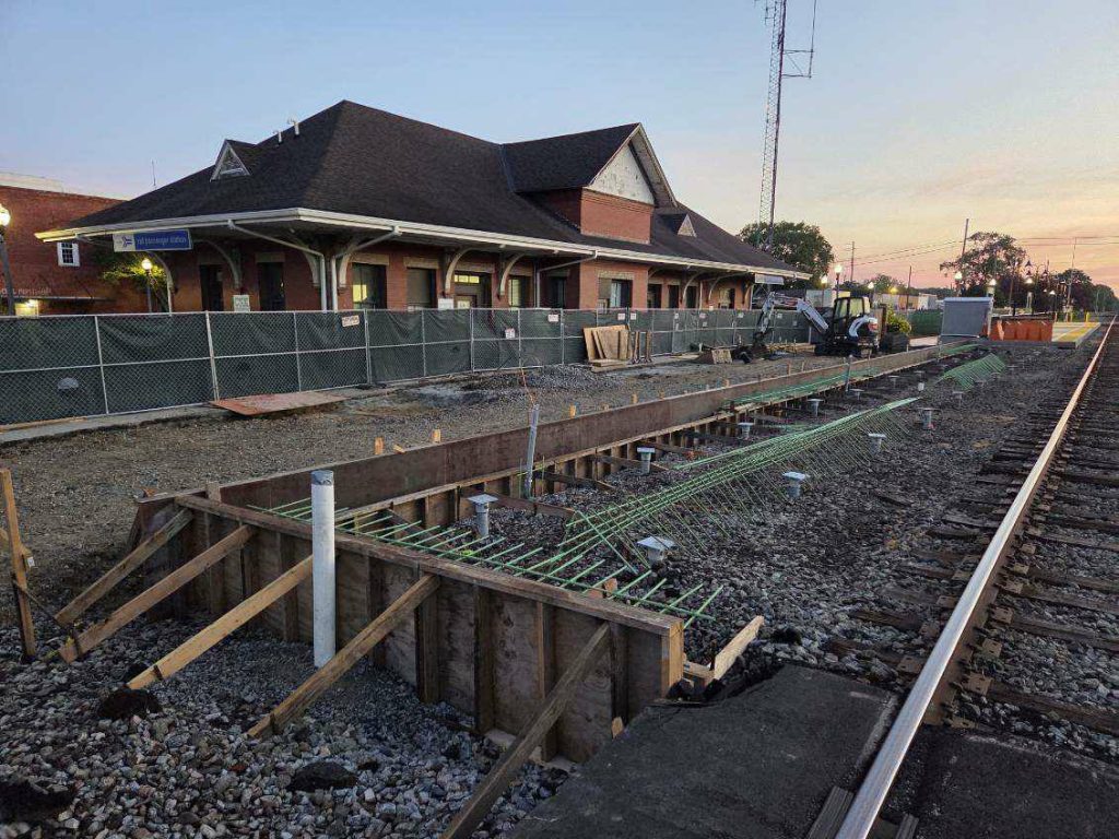 Train station platform under construction beside railroad tracks, fenced off area, construction materials and equipment, brick station building in the background, evening sky.