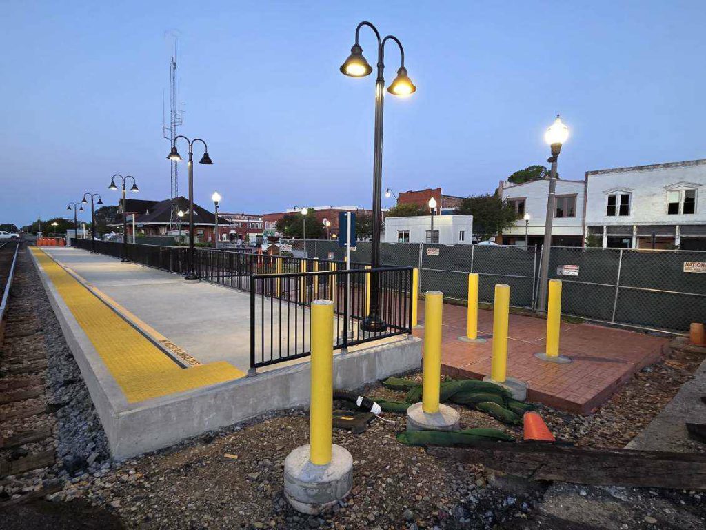 Train platform at dusk with street lamps, yellow safety lines, fencing, and surrounding buildings in a small town setting.