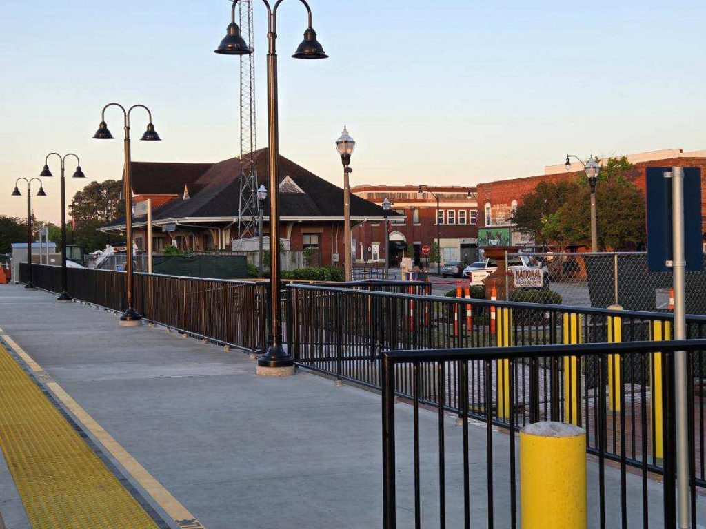 Train station platform with black railings, yellow accessibility strip, street lamps, and a view of brick buildings and parked cars in the background during daylight.