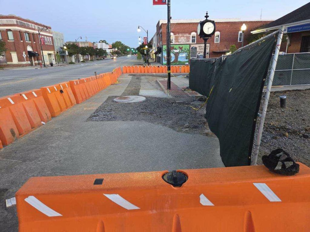 Orange barricades and green fencing block a downtown sidewalk under construction, with a street clock and old brick buildings visible in the background at dusk.