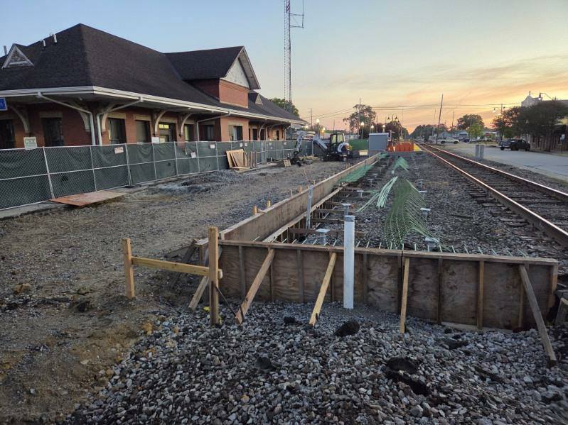 Train station platform under construction beside railroad tracks, with fencing, equipment, and exposed rebar, during sunset.