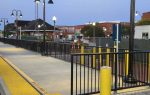 A train station platform with black railings, yellow tactile paving, street lamps, and buildings in the background during early evening.