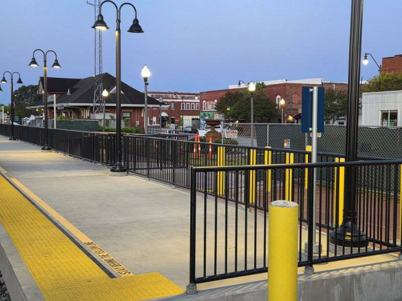 A train station platform with black railings, yellow tactile paving, street lamps, and buildings in the background during early evening.