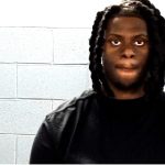 A person with braided hair wearing a black shirt is in front of a white brick wall background.