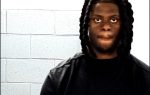 A person with braided hair wearing a black shirt is in front of a white brick wall background.