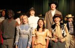 Group of young performers singing on stage, dressed in vintage Western-style costumes with hats and microphones during a theatrical production.