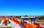 Colorful shipping containers stacked at a busy port terminal with cranes, vehicles, and clear blue sky.