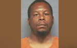 A man in an orange prison uniform facing the camera for a mugshot, with a neutral background.