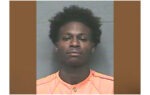 A young man with short curly hair wearing an orange shirt, facing the camera for a mugshot against a grey background.