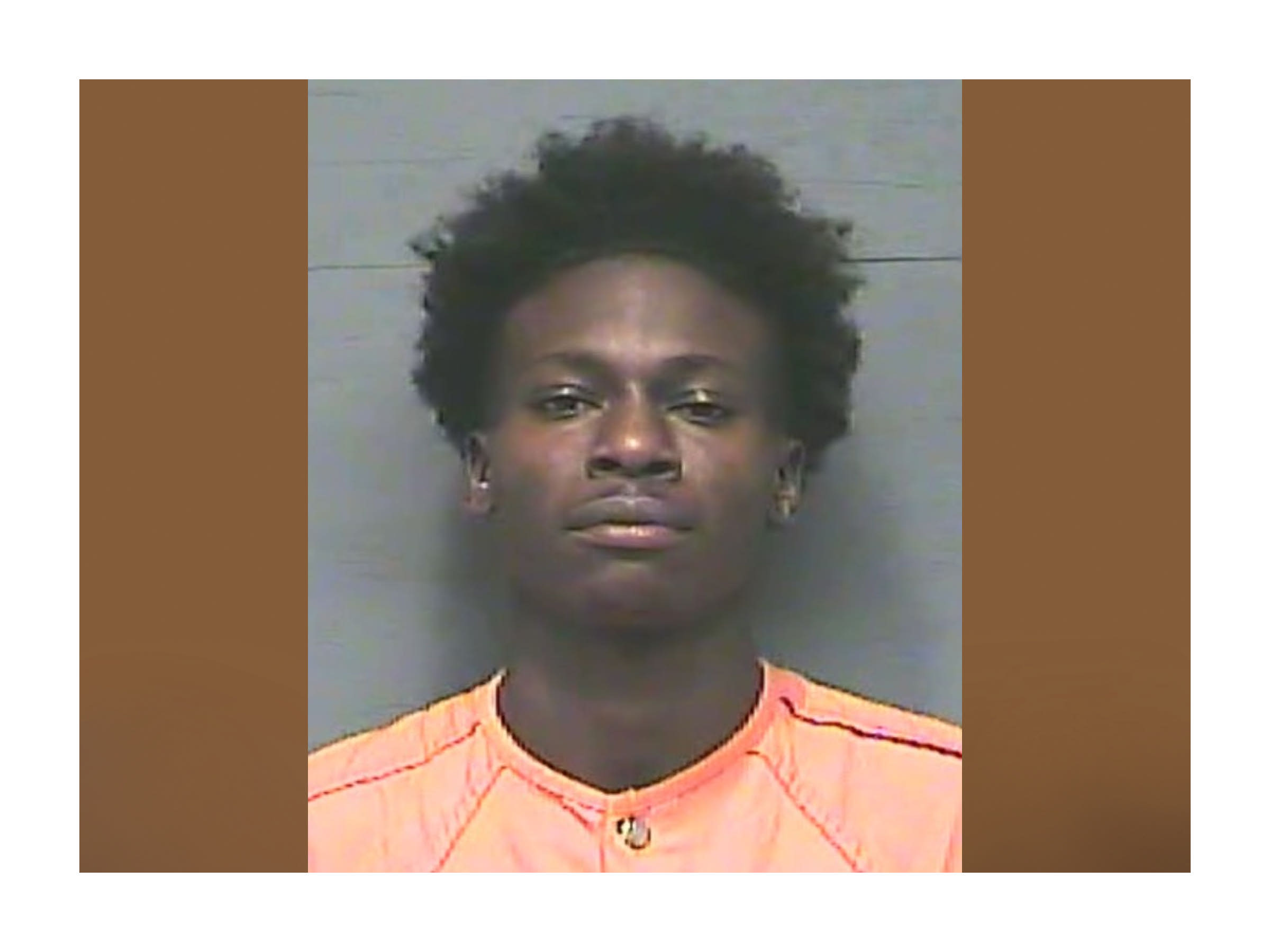 A young man with short curly hair wearing an orange shirt, facing the camera for a mugshot against a grey background.
