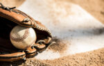 A baseball and glove resting on the infield dirt near home plate, with sunlight casting shadows on the ground.