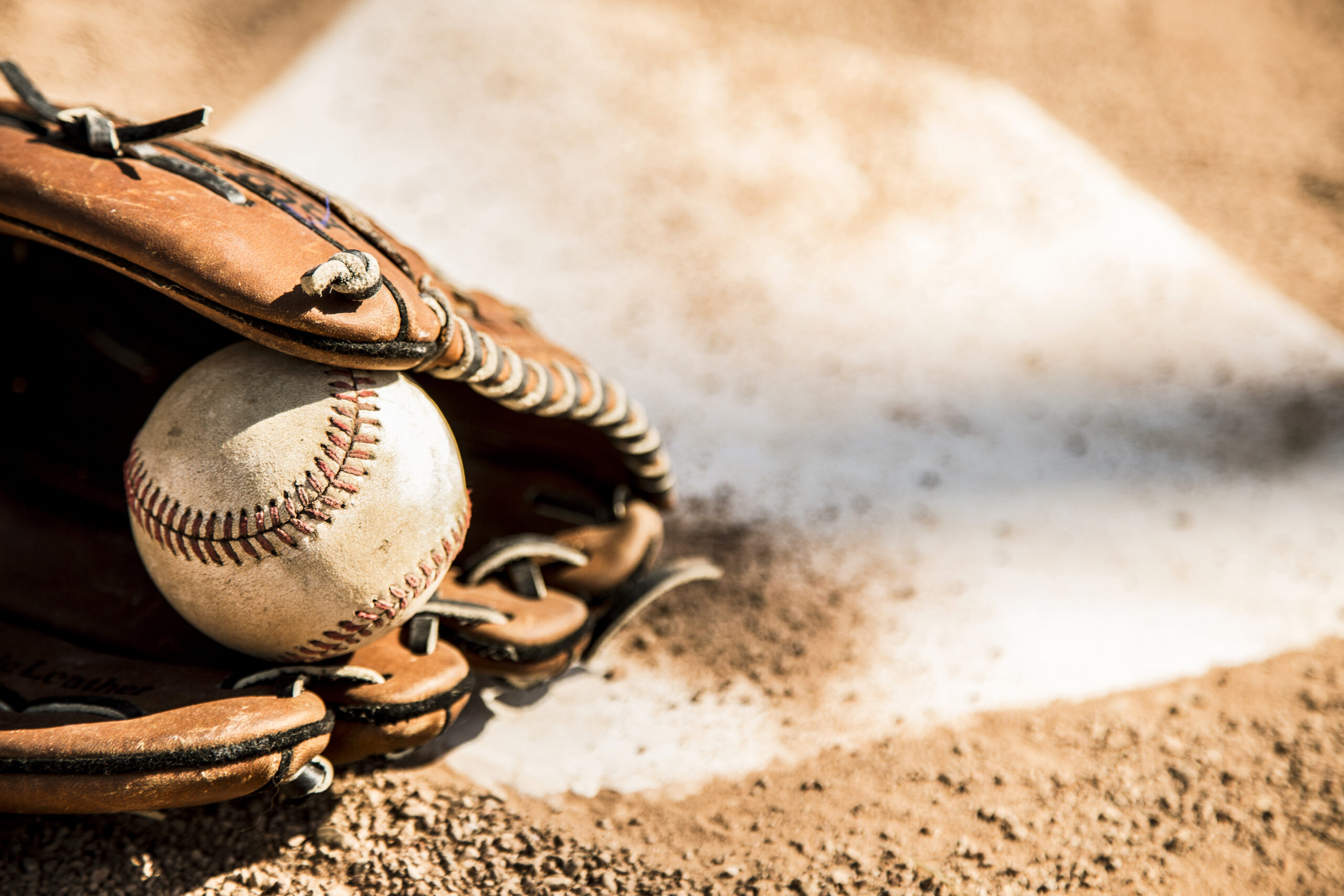 A baseball and glove resting on the infield dirt near home plate, with sunlight casting shadows on the ground.