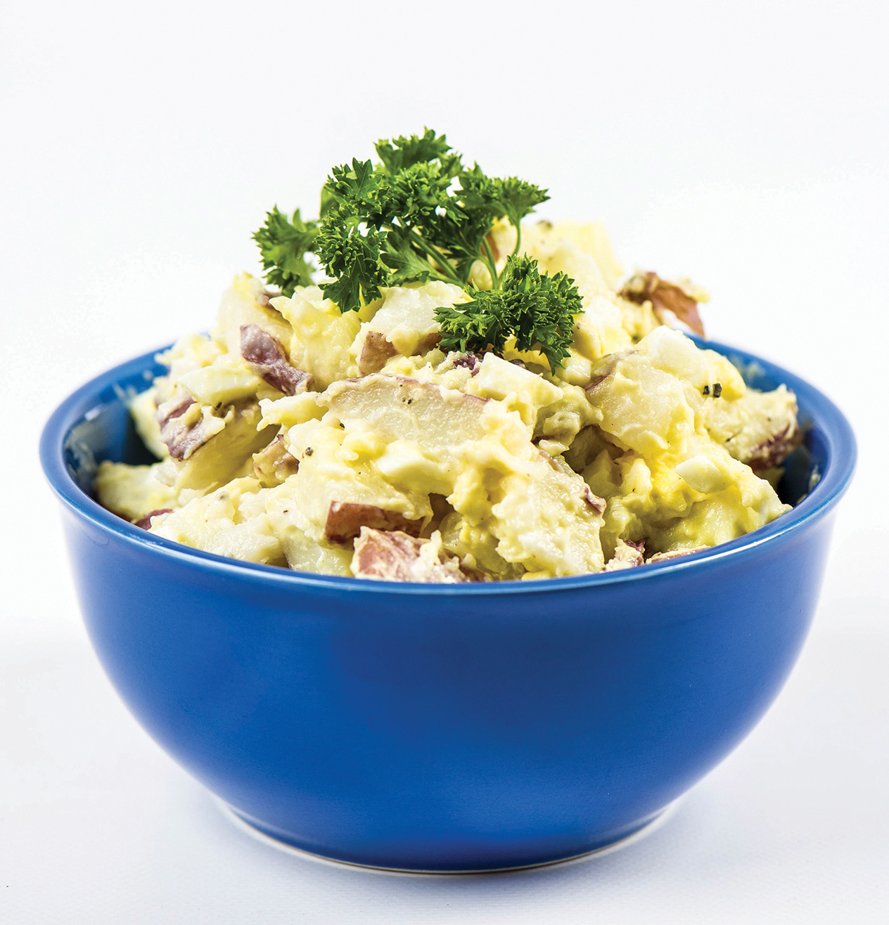 Potato salad with mayonnaise and chopped herbs served in a blue bowl, garnished with a sprig of parsley.