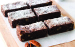 Six rectangular brownies topped with powdered sugar are arranged on a wooden cutting board, with pecans beside them and a small bowl of pecans in the background.