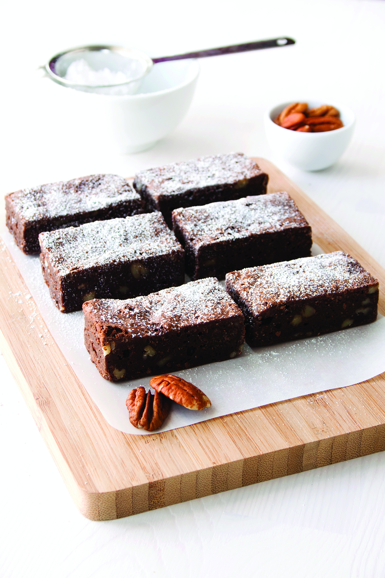 Six rectangular brownies topped with powdered sugar are arranged on a wooden cutting board, with pecans beside them and a small bowl of pecans in the background.