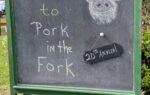 Chalkboard sign reads "Welcome to Pork in the Fork 21st Annual" with a drawing of a pig wearing glasses, displayed outdoors on grass.