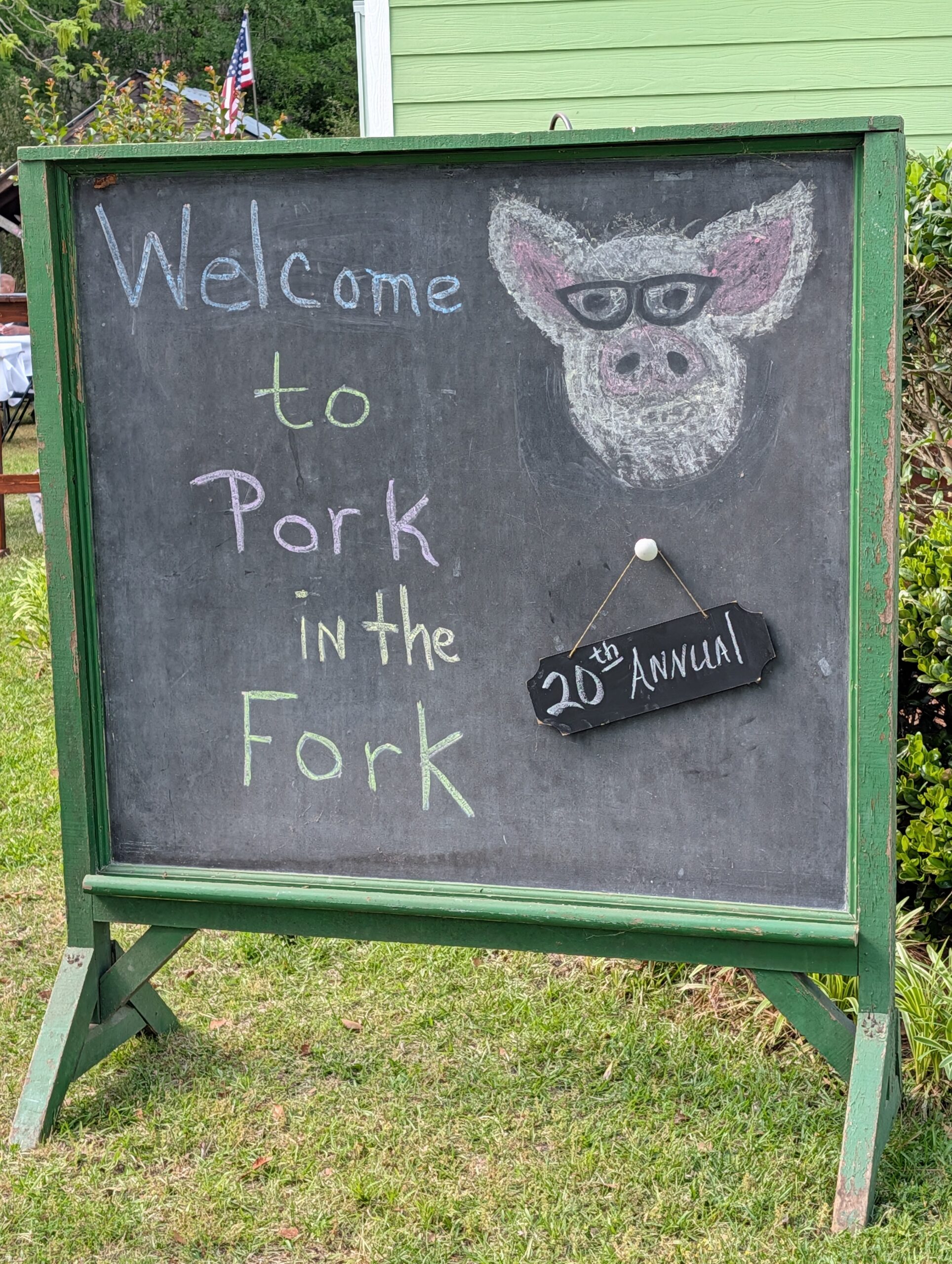 Chalkboard sign reads "Welcome to Pork in the Fork 21st Annual" with a drawing of a pig wearing glasses, displayed outdoors on grass.