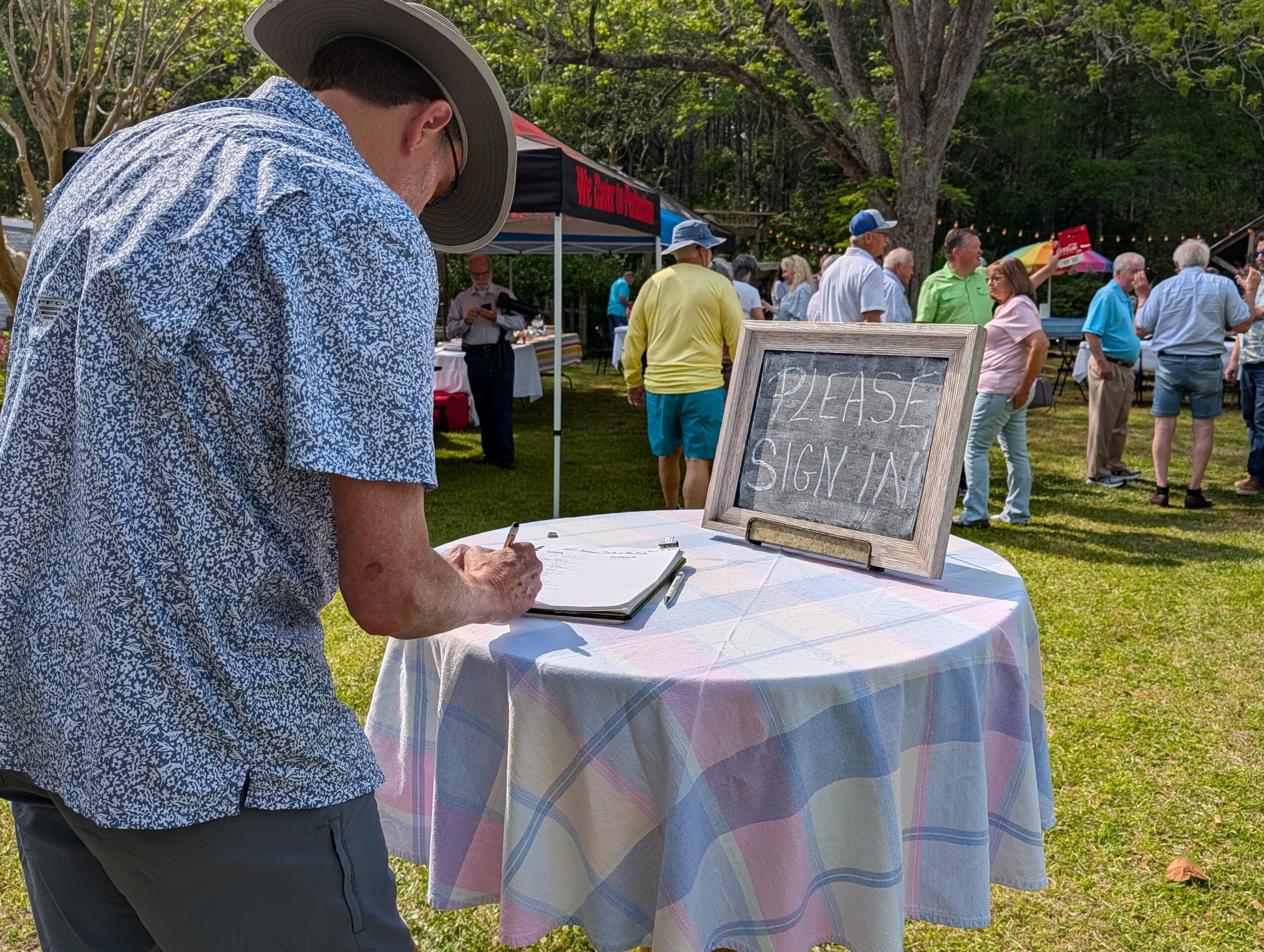 A person wearing a hat writes on a clipboard at a round table outdoors during a community event, with other people and tents visible in the background.