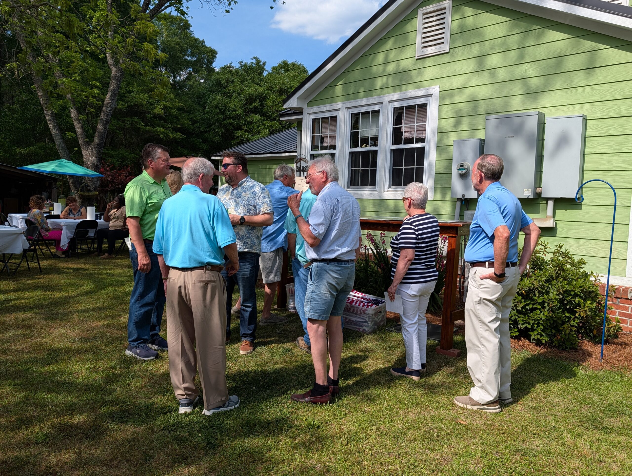 A group of older adults standing and talking outside a green house on a sunny day, with trees and clear sky in the background.