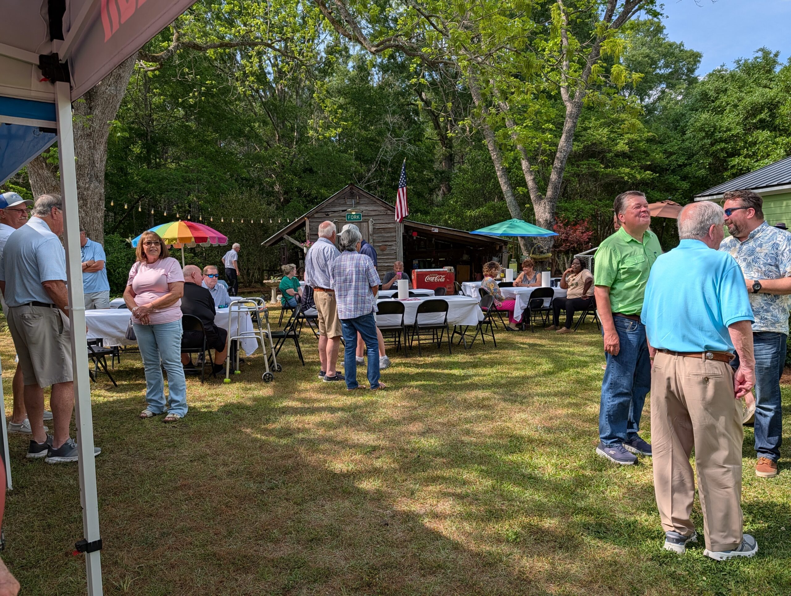People gathered outdoors at a picnic with tables, chairs, and colorful umbrellas, surrounded by trees on a sunny day.