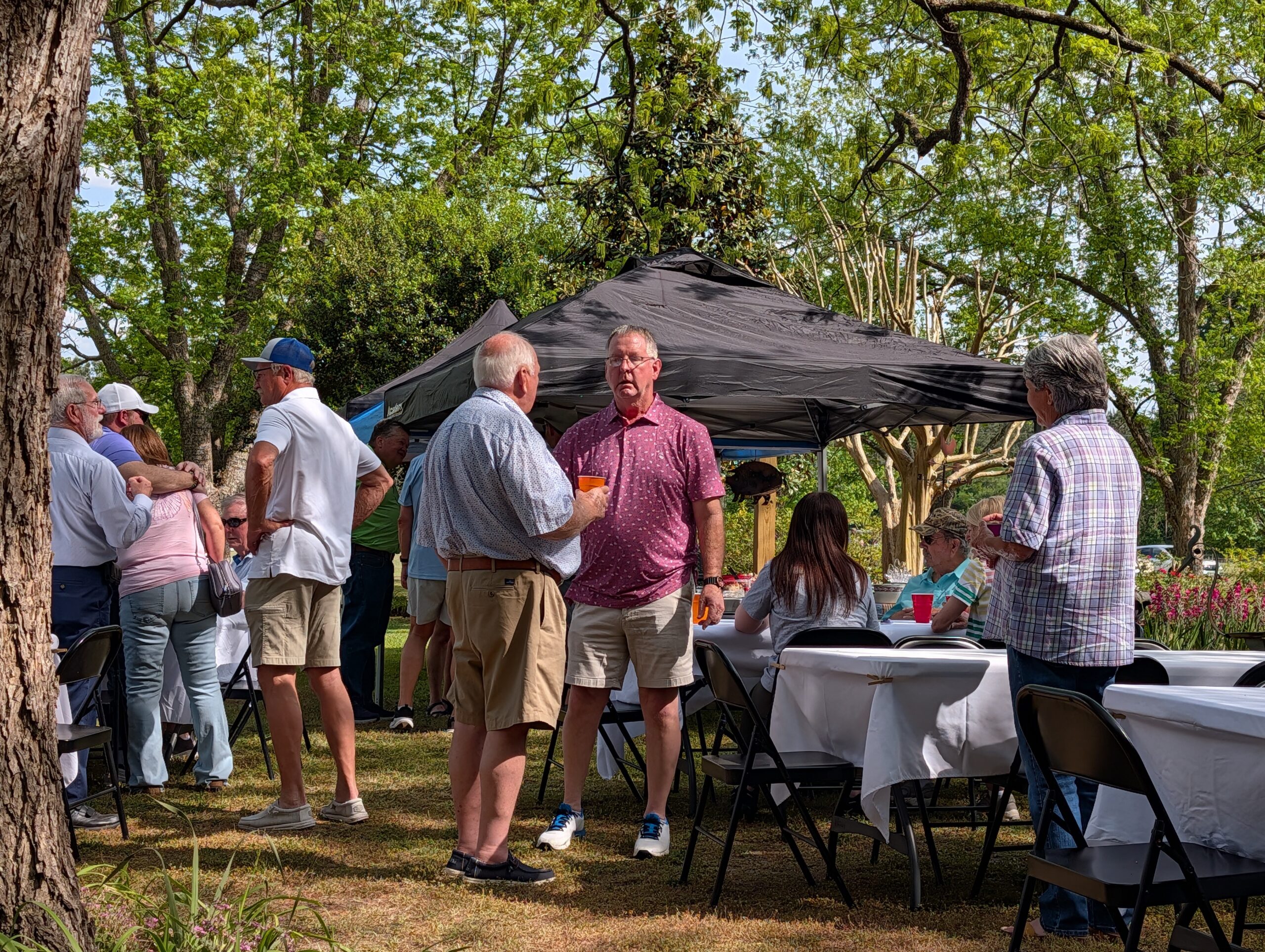 People socializing outdoors at a garden party with tables, chairs, and a canopy tent, surrounded by trees and greenery on a sunny day.