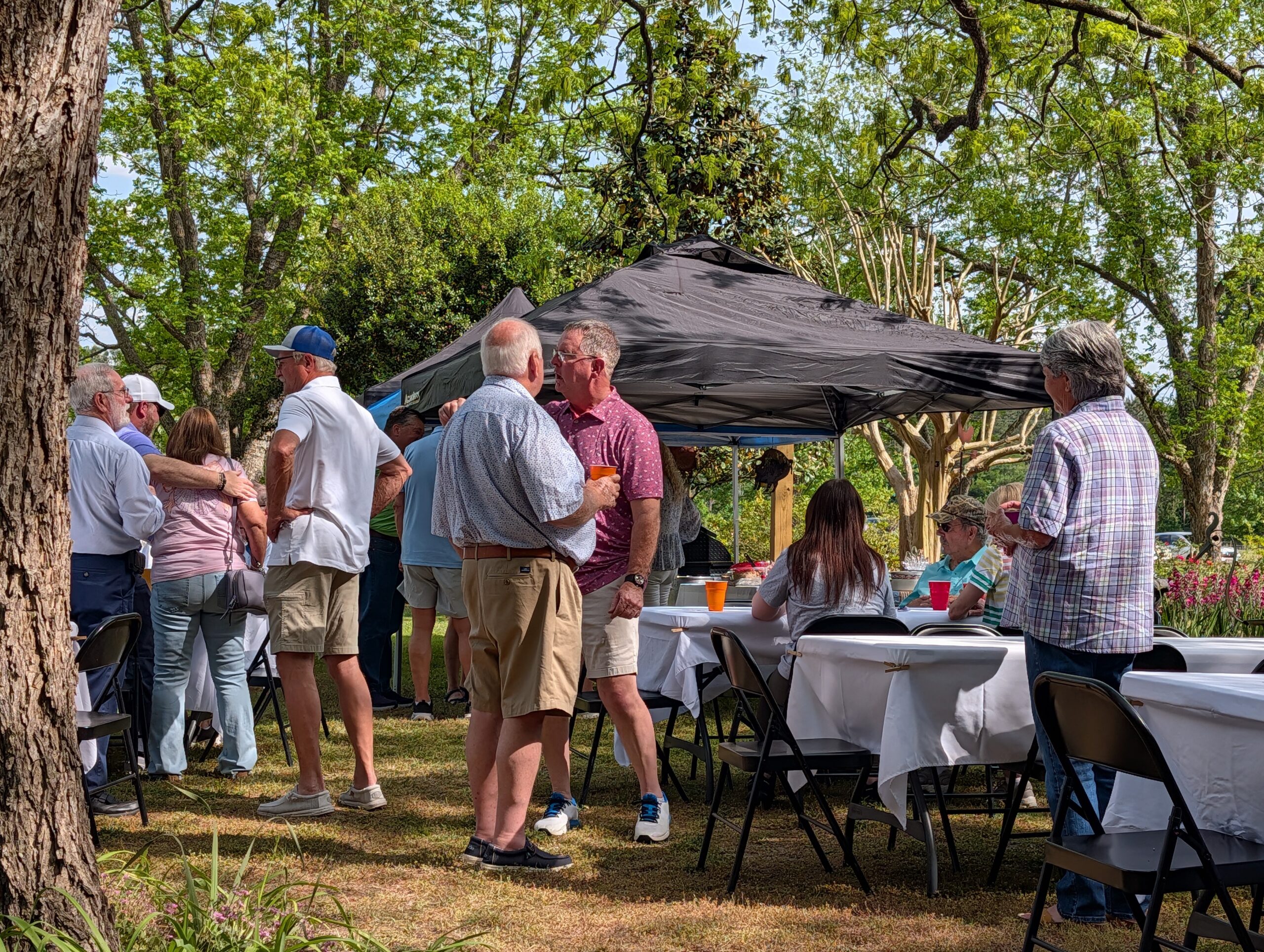People gather outdoors under trees, talking and eating at tables covered with white cloths, with a black canopy providing shade on a sunny day.