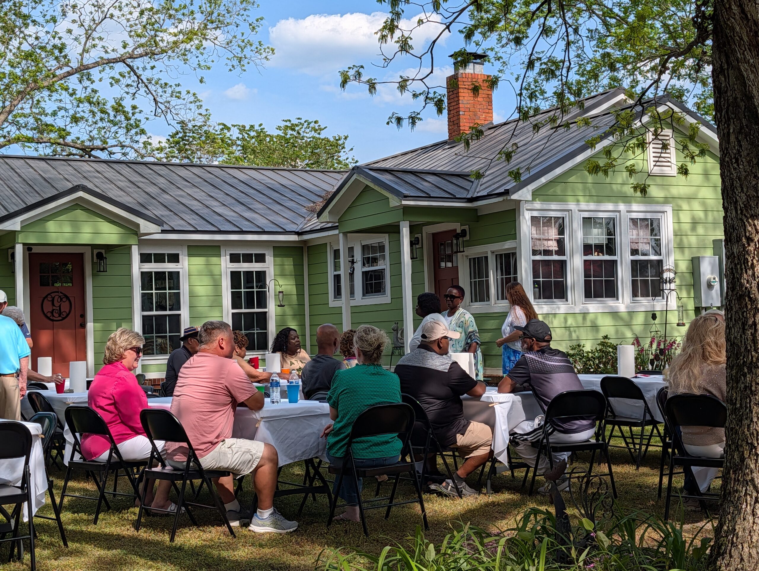 People gathered outdoors around tables in a backyard, enjoying a meal together on a sunny day near a green house.