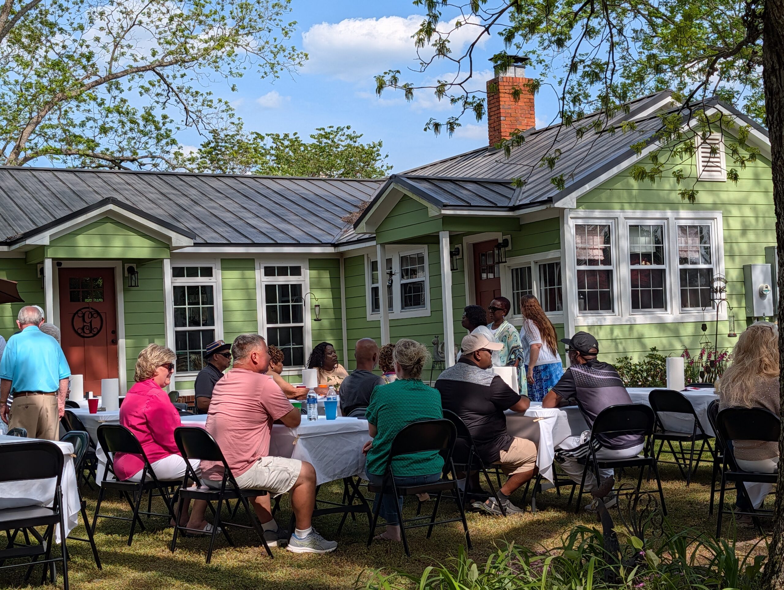 People sitting at tables outside a green house, enjoying a gathering on a sunny day with trees and blue sky in the background.