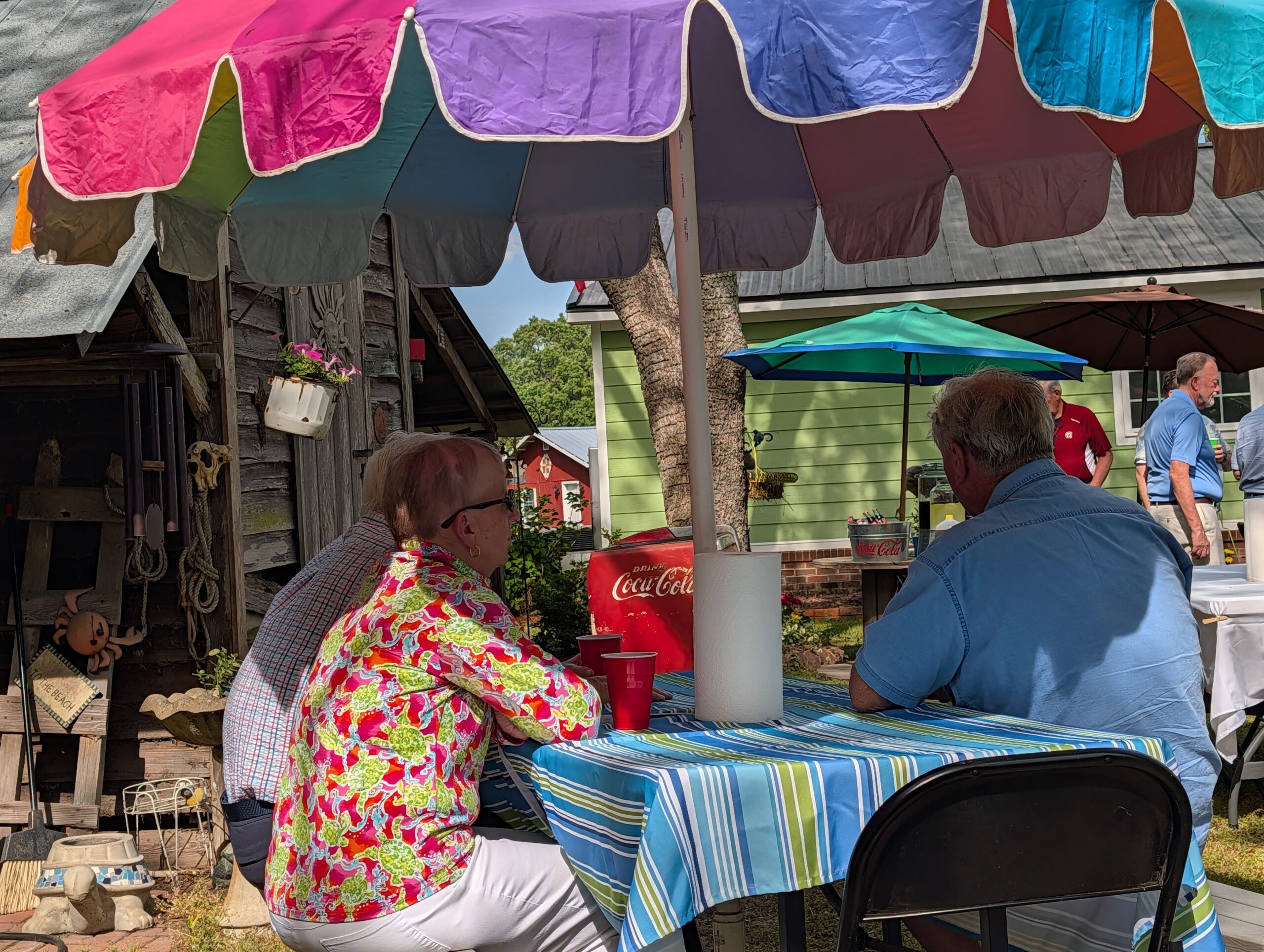 People sitting at a table outdoors under a colorful canopy, with a house and trees in the background. A paper towel roll and drinks are on the table.