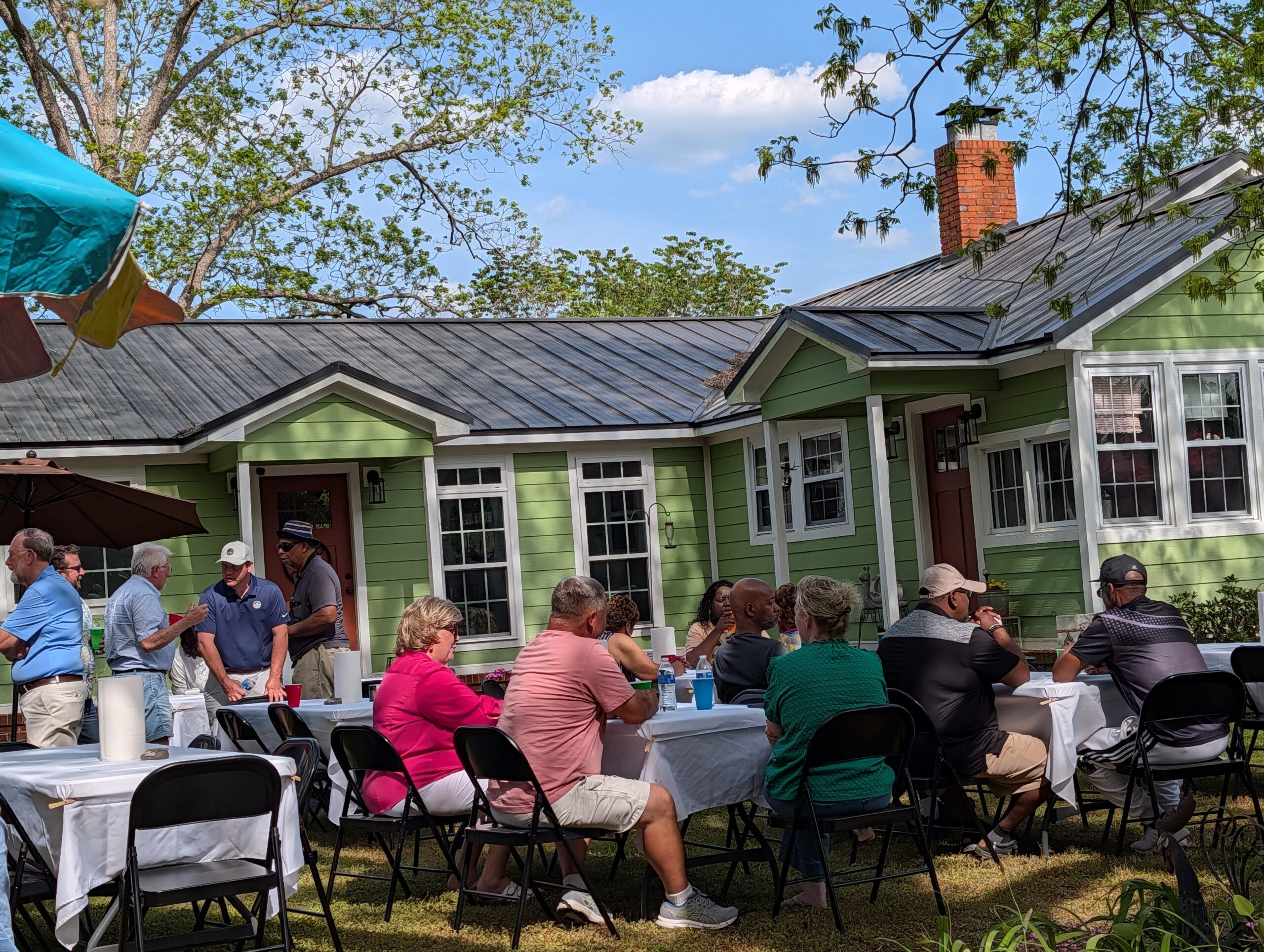 People gathered outside a green house, sitting at tables with white tablecloths, enjoying a meal under a sunny sky with trees and blue clouds in the background.