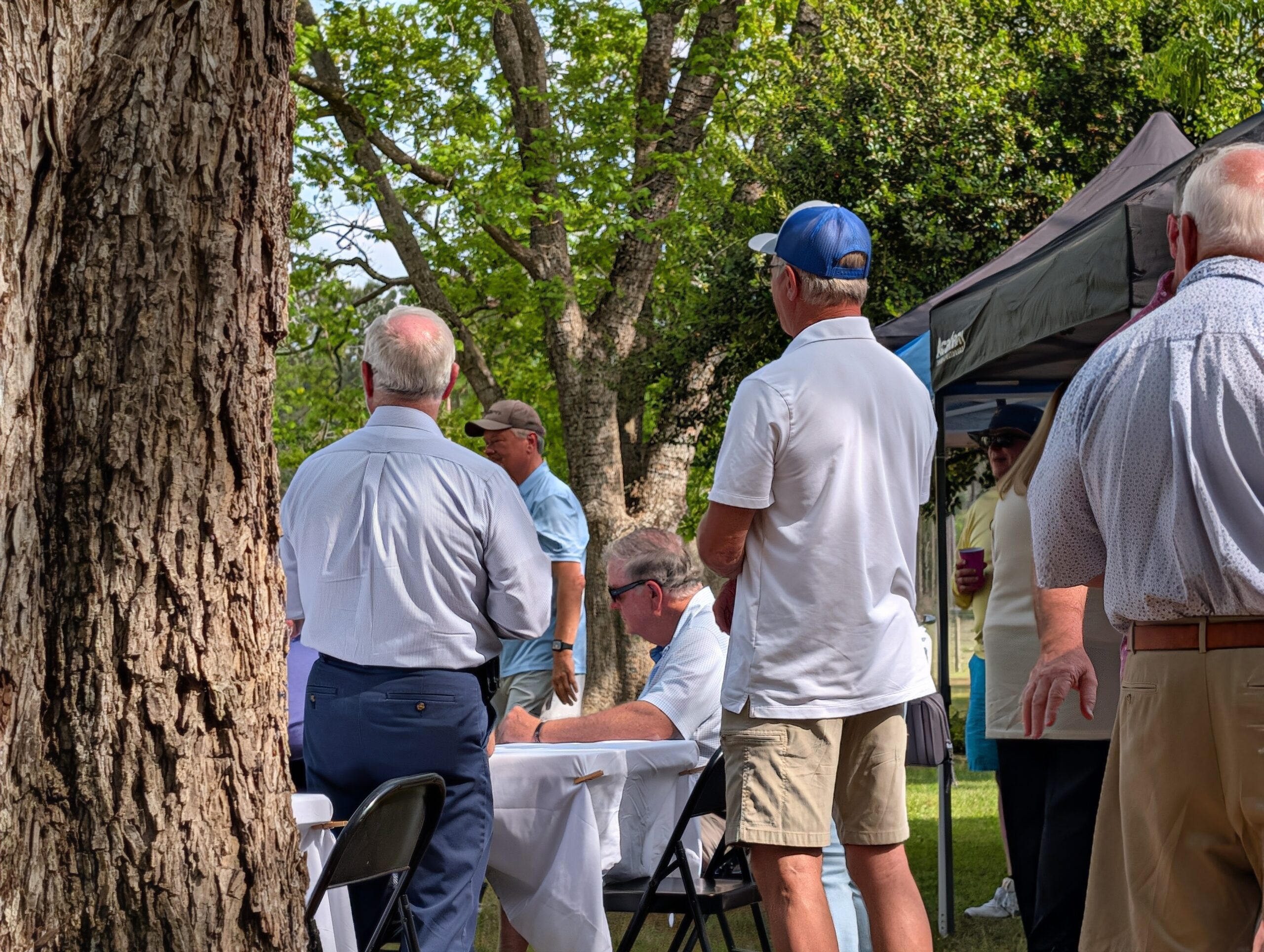 People gathered outdoors near tables and a canopy, some standing, some seated, surrounded by trees on a sunny day.