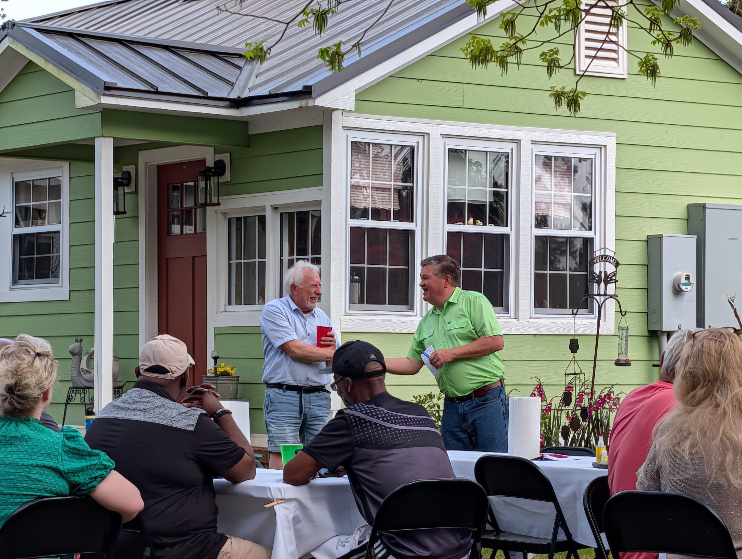 People gathered outside a green house, some sitting at a table and two men standing and talking near the entrance.