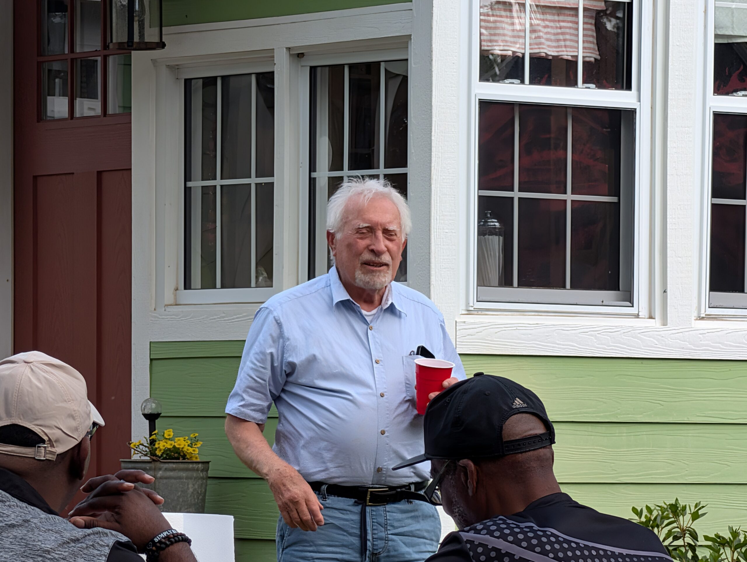 older man with white hair stands outside a green house, holding a red cup. Several people are seated in the foreground.