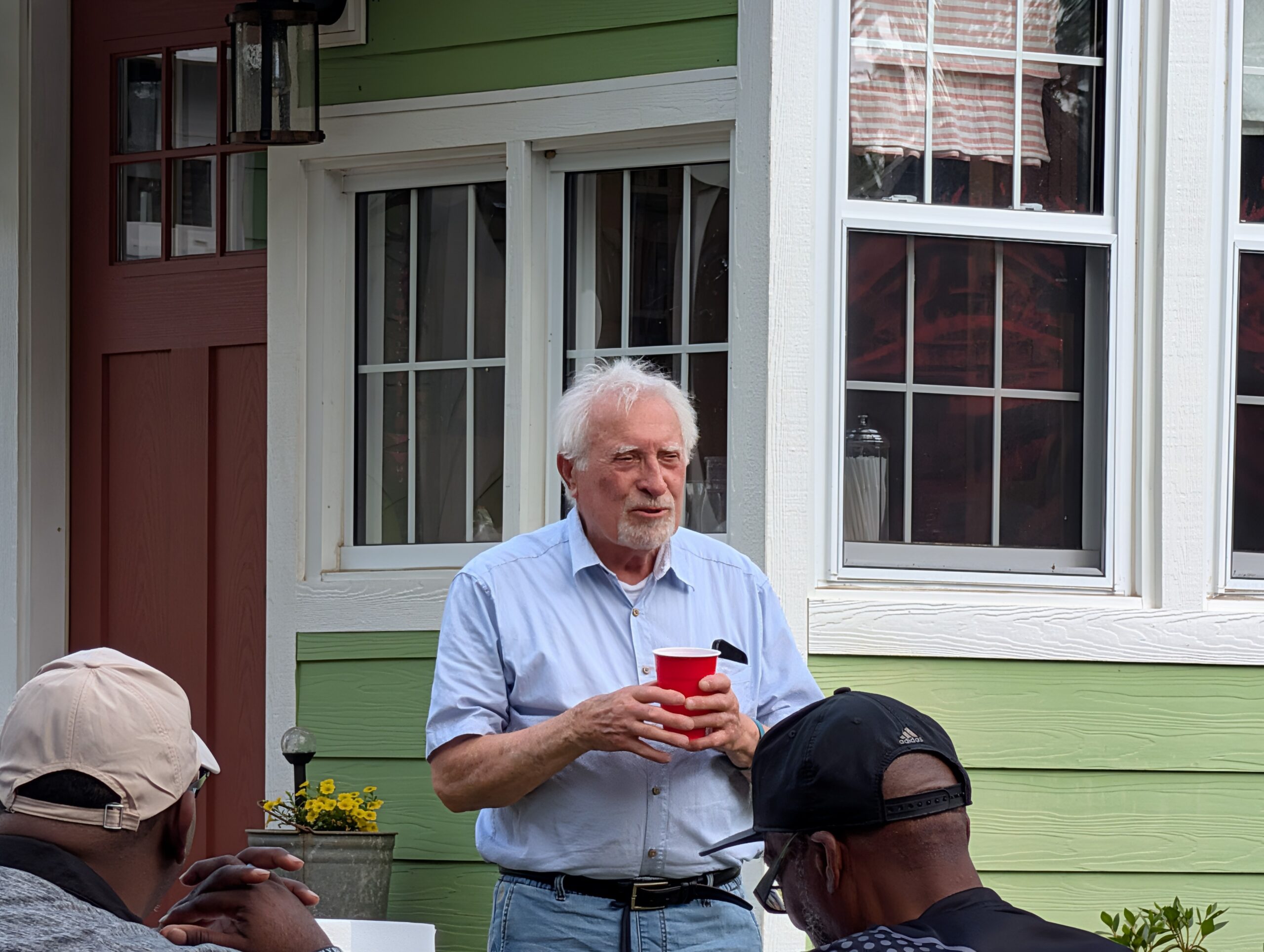 elderly man holding a red cup stands outside near a green house, talking to people seated nearby on a sunny day.