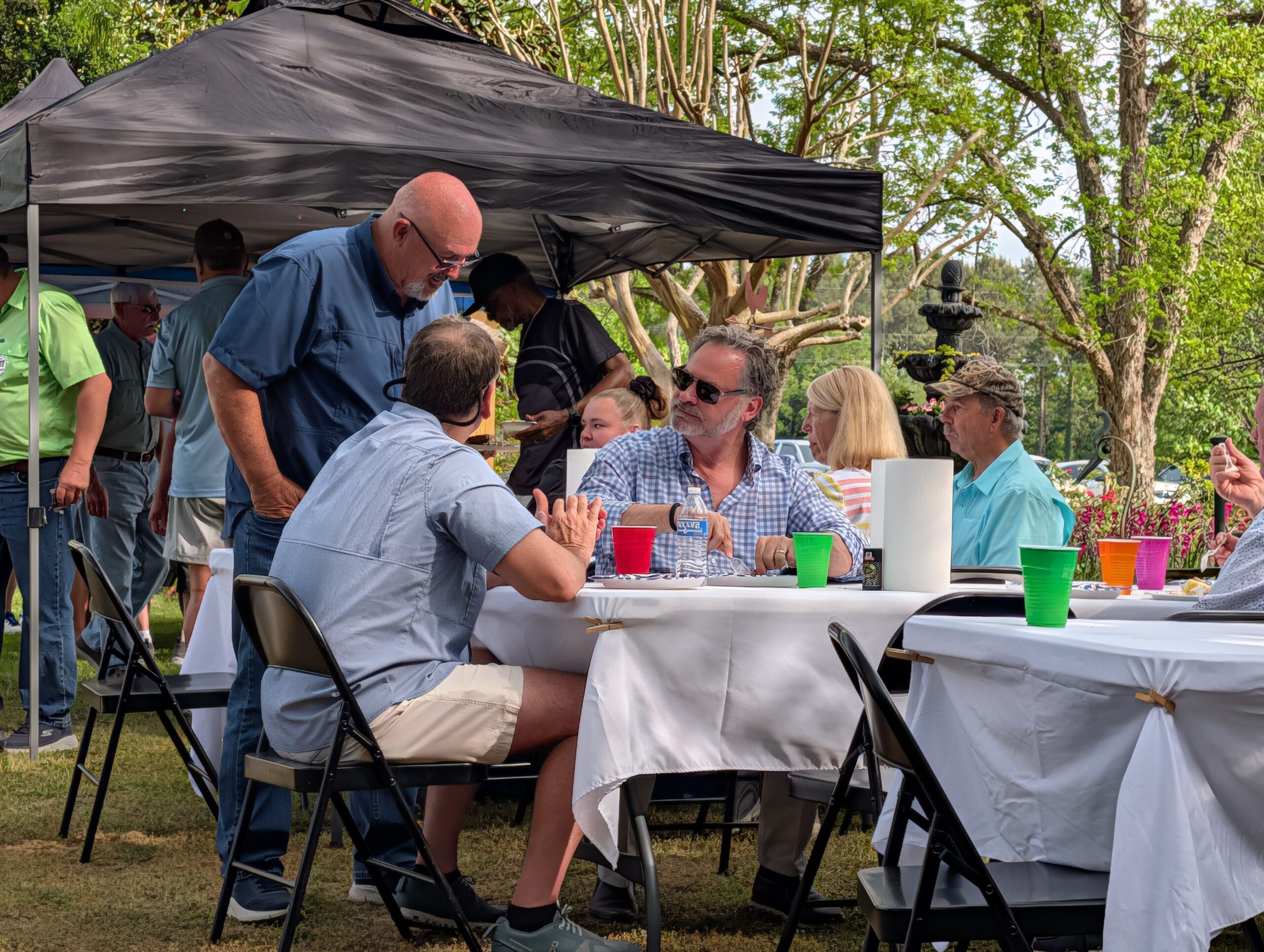 People sitting outdoors at a picnic table covered with a white tablecloth, talking and enjoying food and drinks under a canopy on a sunny day.