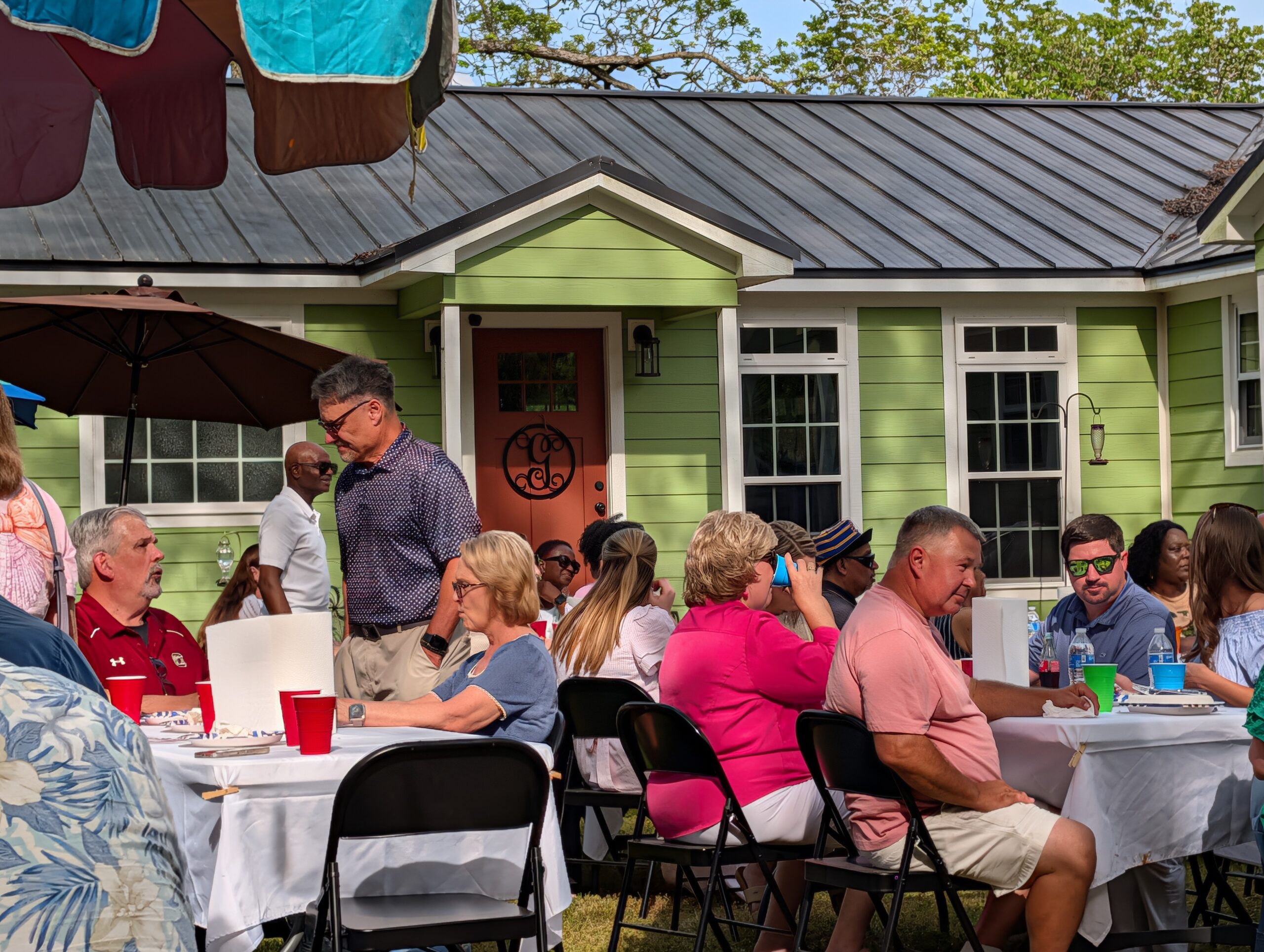 People gathered outside a green house, sitting at tables with food and drinks, enjoying a casual outdoor social event on a sunny day.