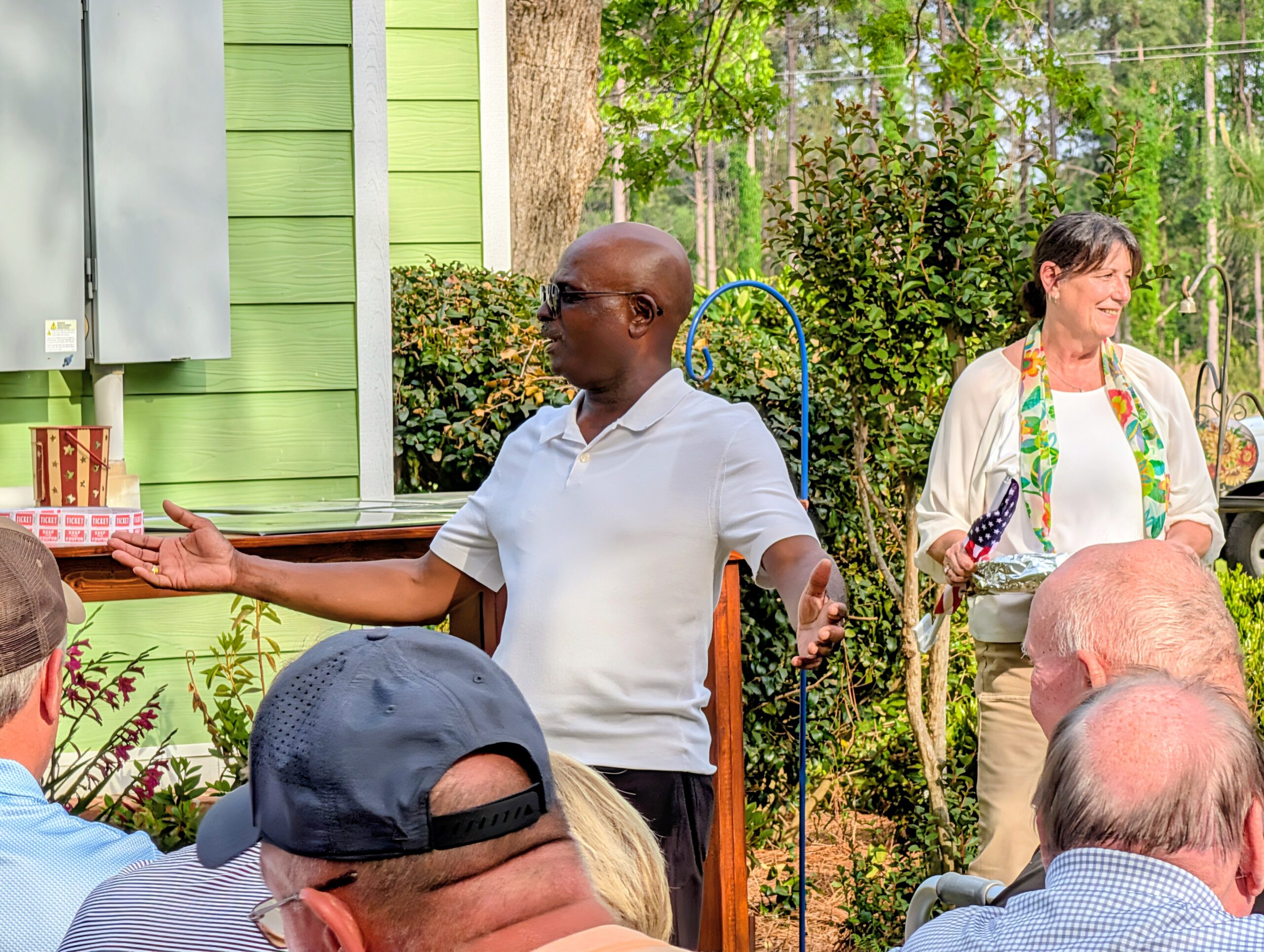 A man in a white shirt speaks outdoors to a small group of people; greenery and a light green house are in the background.