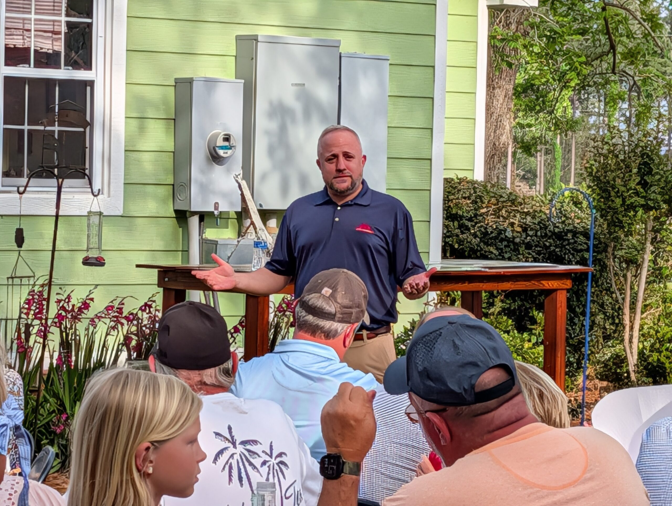 A man speaks to a small group of people outdoors by a green house, gesturing with his hands during a presentation or meeting.