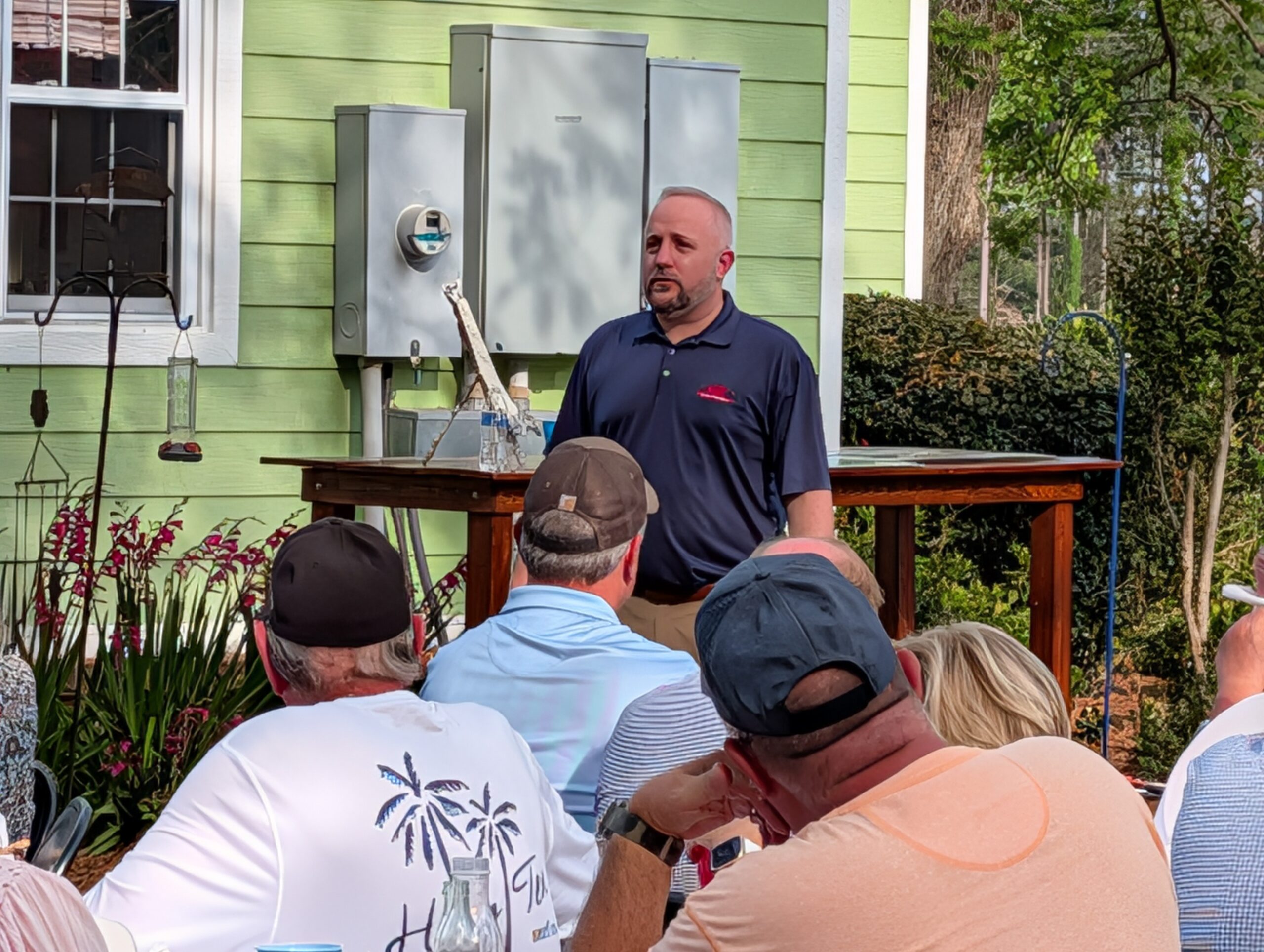 A man stands and speaks to a seated group outdoors near a green house with utility boxes on the wall.