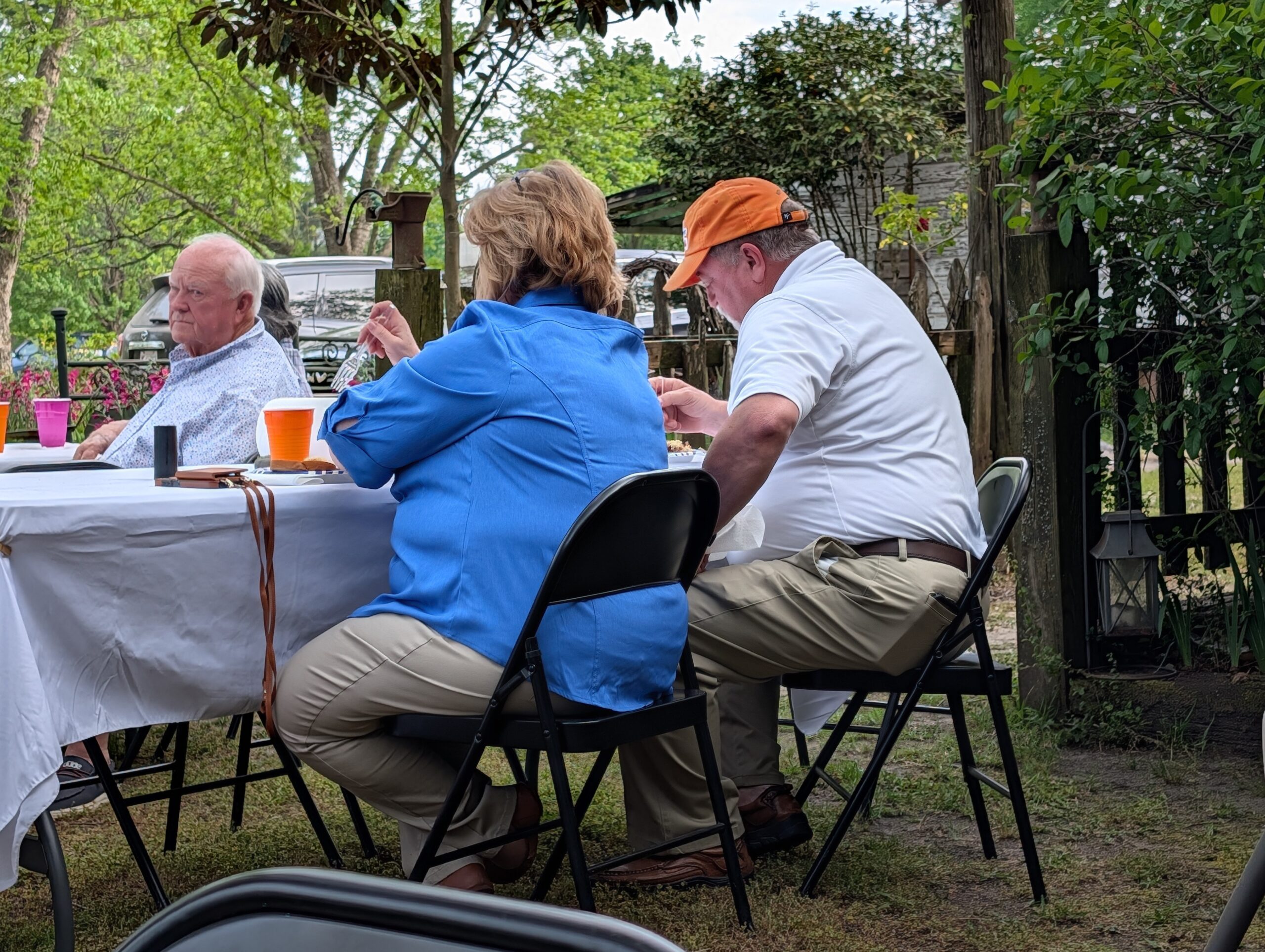 Three older adults sit at an outdoor table covered with a white tablecloth, eating and talking, surrounded by trees and greenery in a backyard setting.
