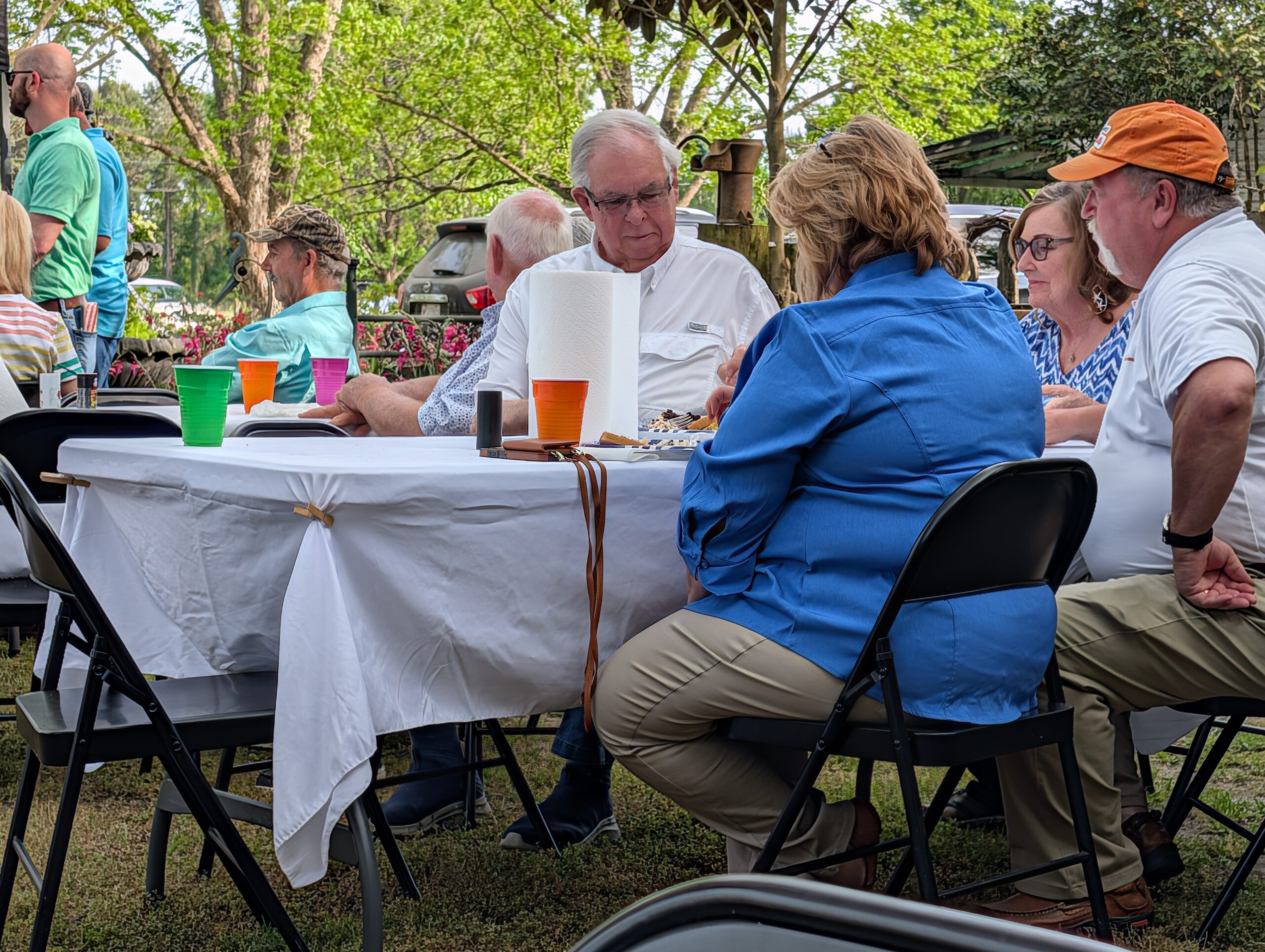 A group of people sitting around a table outdoors, eating and talking, with colorful cups and trees in the background.