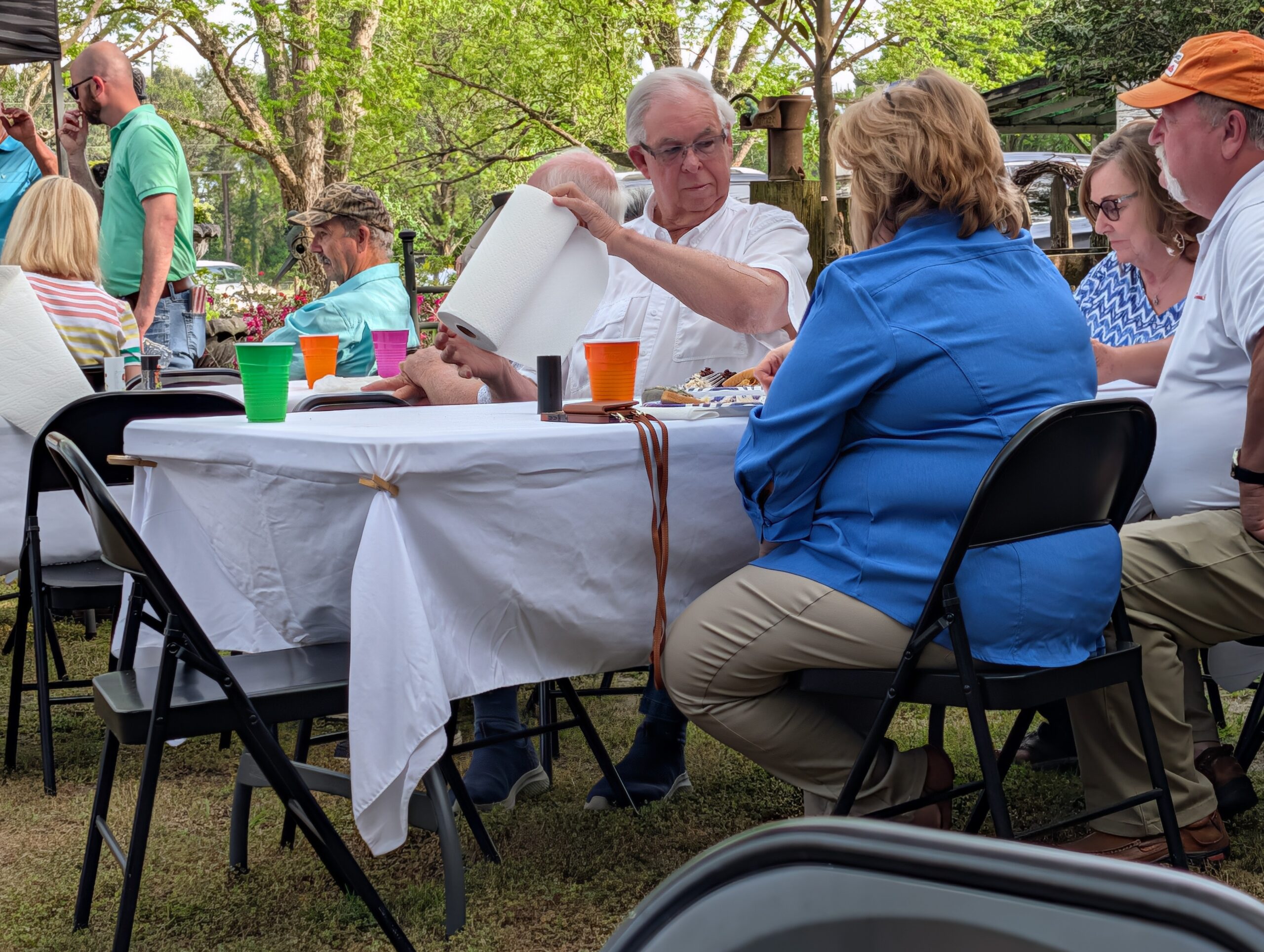 People sitting around a table outdoors, enjoying a meal together; one man is pouring a drink while others converse. Trees and picnic items visible in the background.