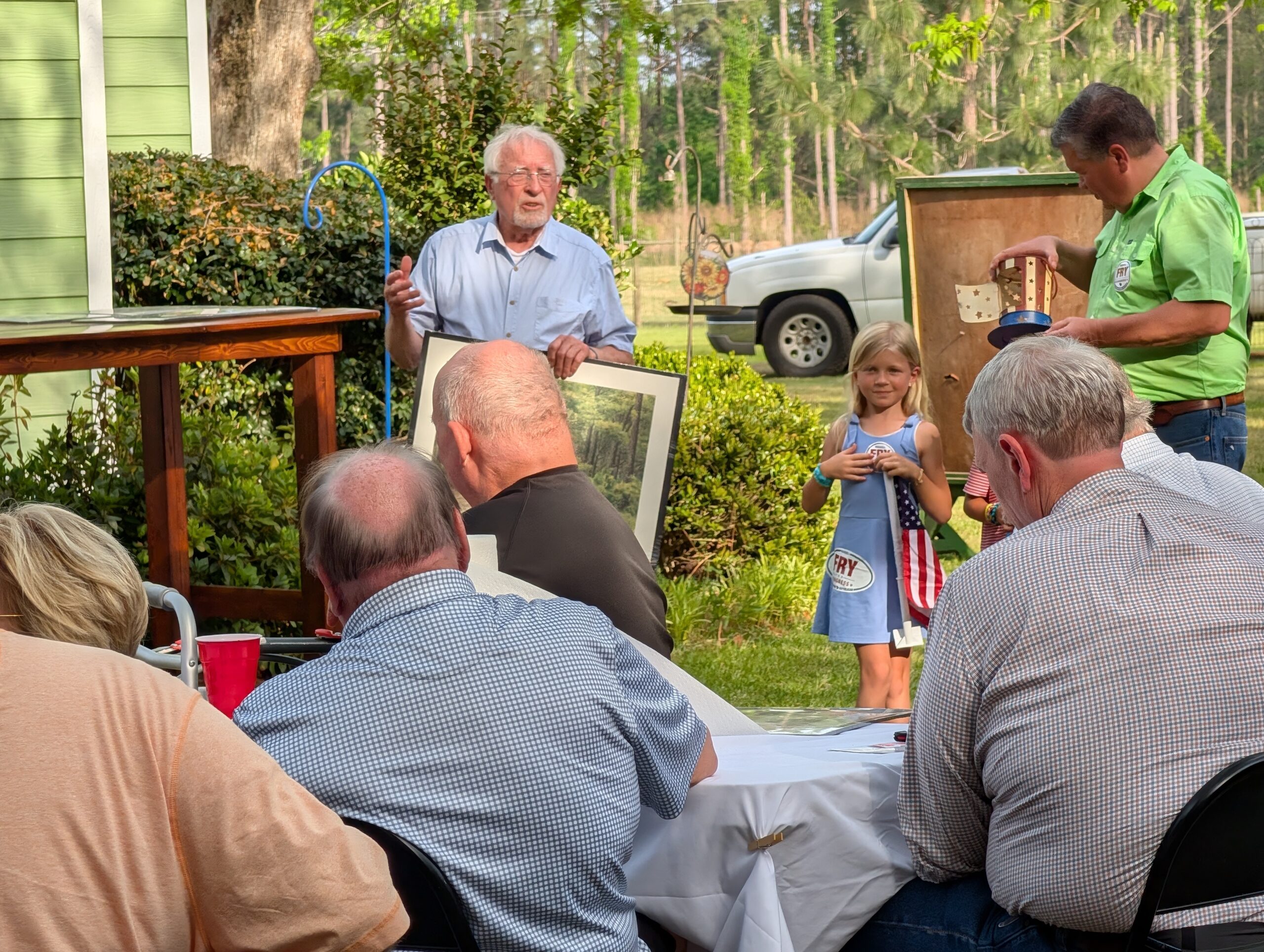 elderly man speaks to a group of people seated outdoors, with a child and other adults nearby, surrounded by trees and parked vehicle in the background.