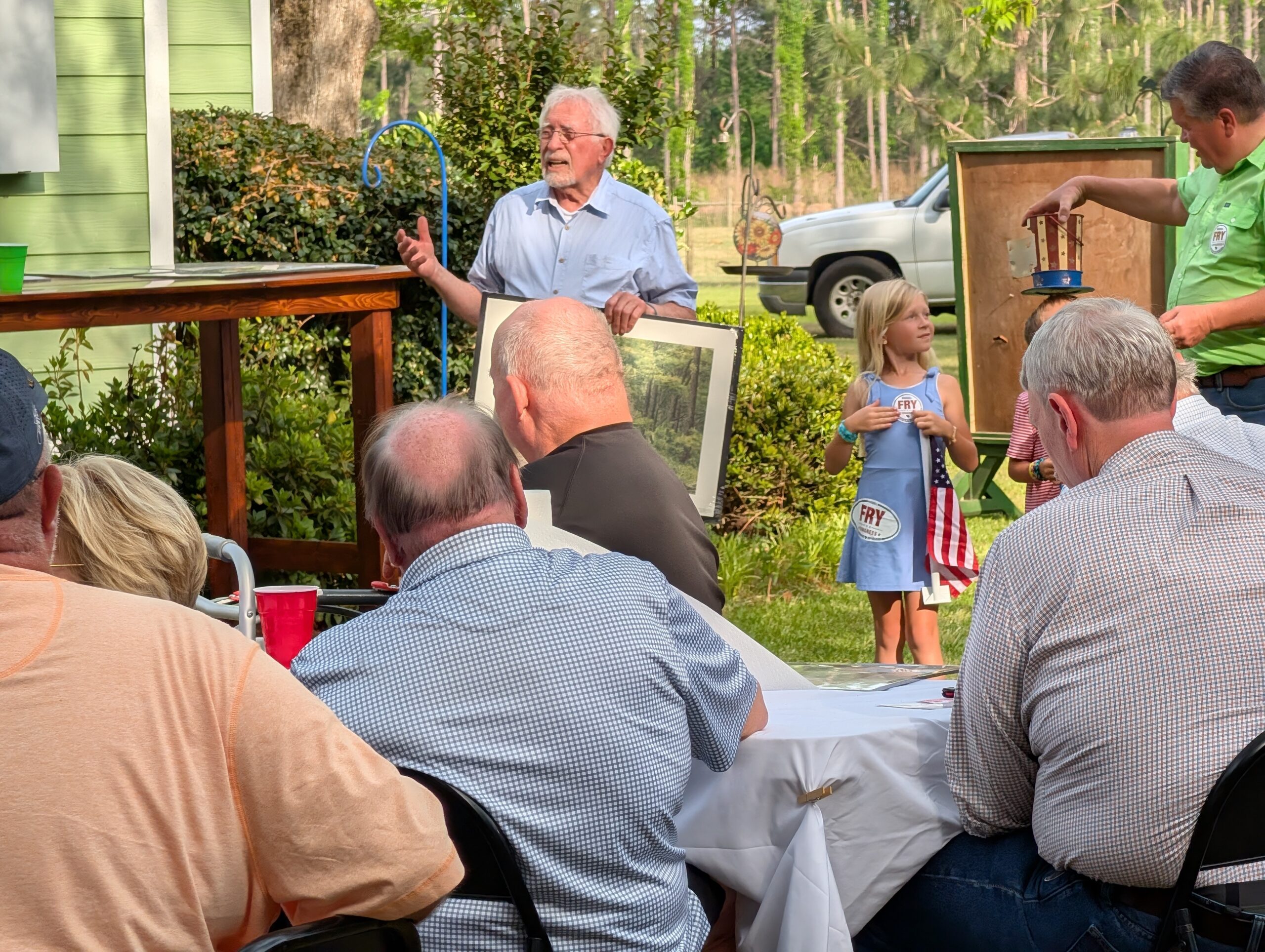 older man speaks to a seated group outdoors while holding a picture frame. A young girl stands beside him, holding an American flag.
