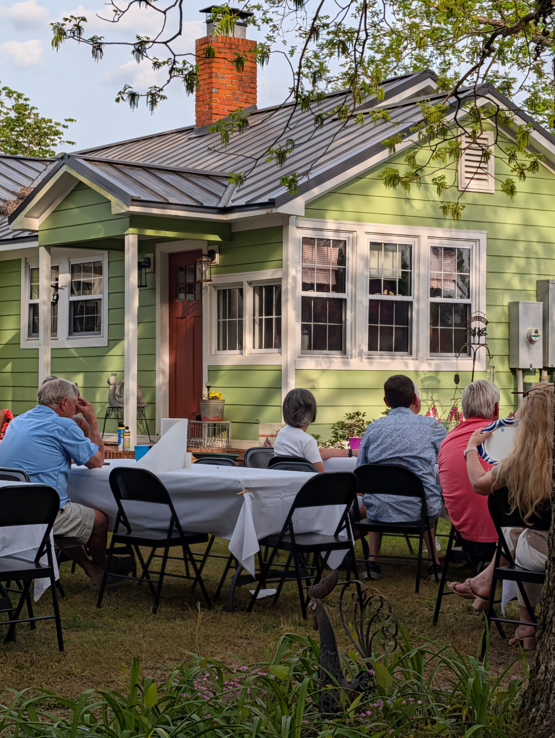 People sit around a long table outdoors in front of a green house, enjoying a gathering or meal together on a sunny day.