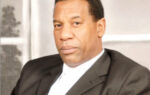 A man in a dark suit and white shirt sits indoors, looking at the camera with a neutral expression.